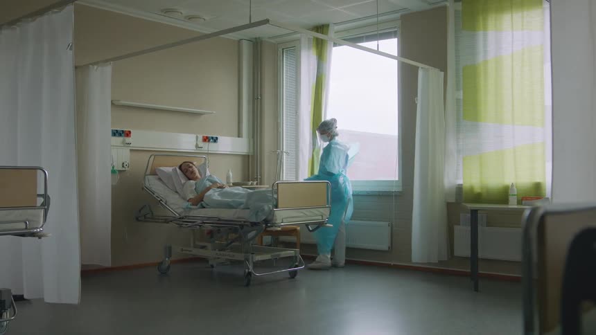 A woman lies in a hospital bed as a nurse walks in the room and pulls open the drapes and goes to the bed to talk with her