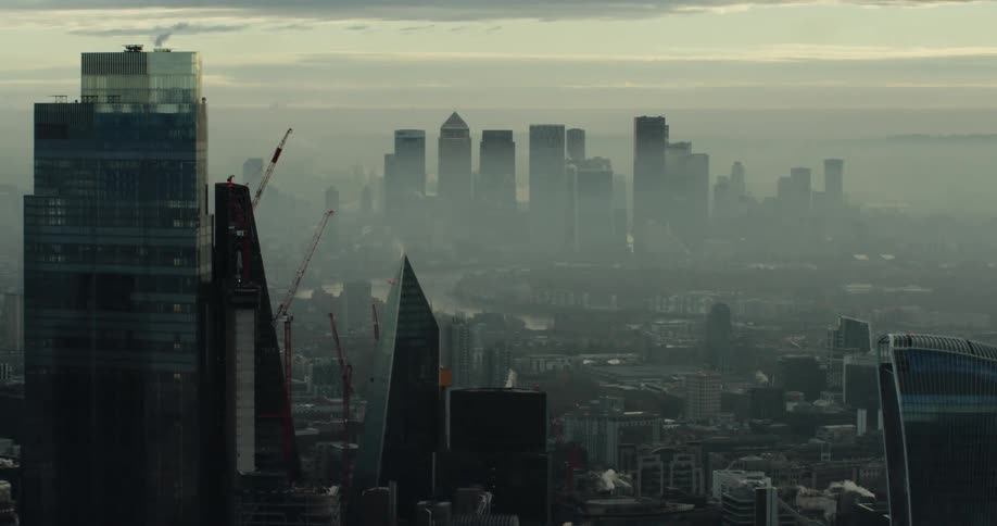Tall city buildings and skyscrapers are seen through the fog under a cloudy sky