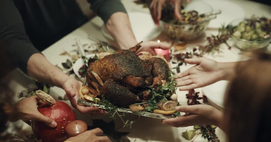 A woman is carrying a thanksgiving turkey to a dinner table where friends are sitting