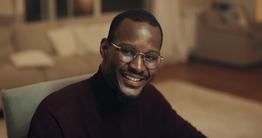 A man who is sitting in a chair in a living room looks to his right and smiles