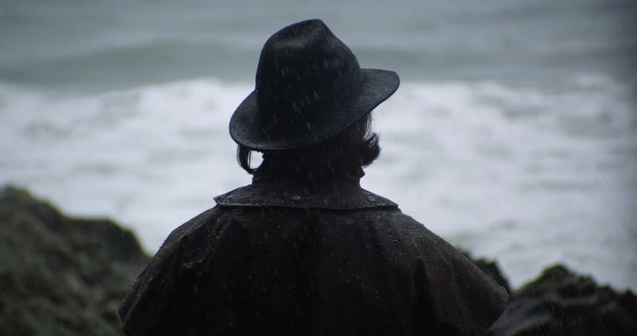 A person wearing a black hat is sitting on rocks along the shore and watching the waves in the rain