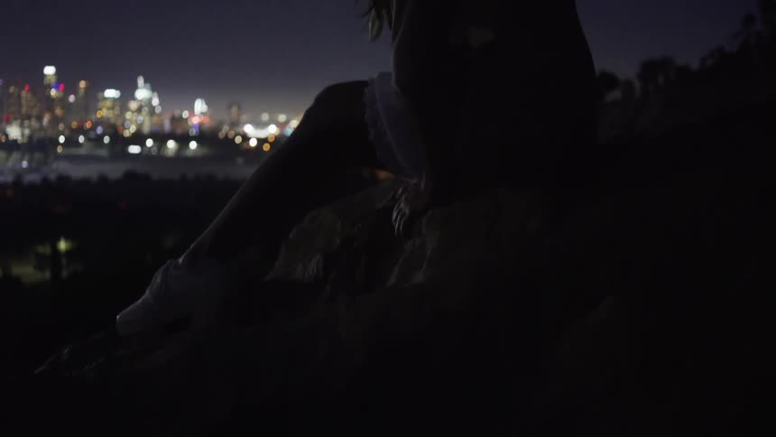 A woman is sitting on a hill top at night overlooking a city