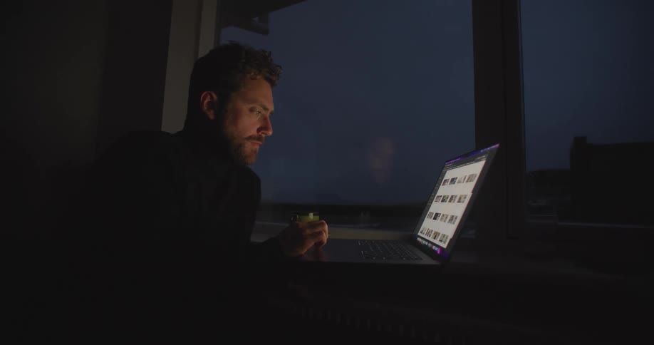 Man types on laptop sitting near window at night