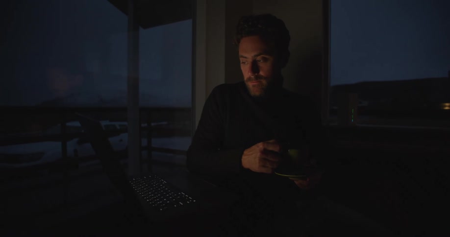 A man is sitting at a table in a dark room drinking from a cup and looking at a laptop computer