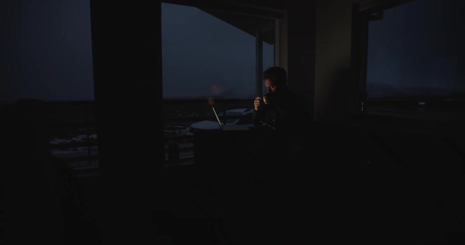Man with beard drinks from cup and works on laptop in dark room at night
