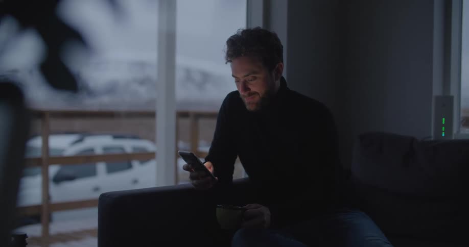 Man sitting on couch next to a window texting on a cell phone