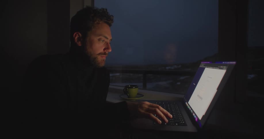 A man is sitting at a table in a dark room typing on a laptop computer