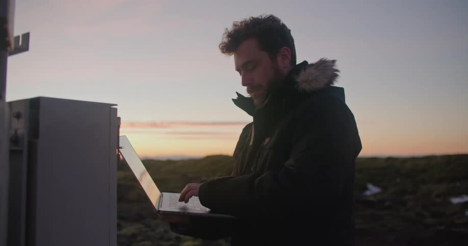 A man is standing outside in front of a utility box holding a laptop computer and typing with the other hand