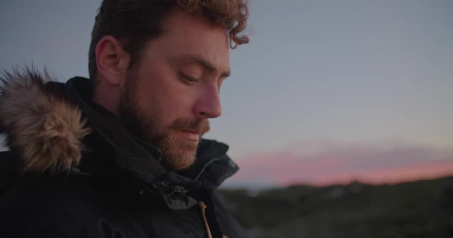 Man with beard and black coat uses laptop outdoors at sunset