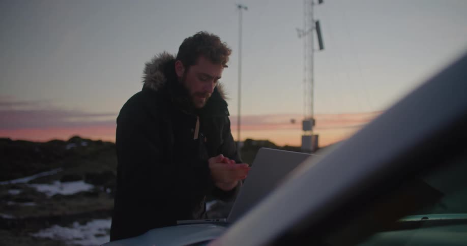 A man is standing outside wearing a winter coat and typing on a laptop computer that is sitting on the hood of a car with utility towers nearby