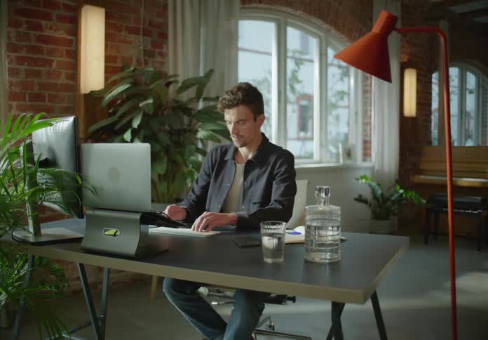 Man with goatee with black shirt and jeans sits at desk working on laptop in room