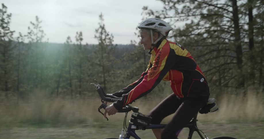 Older female bicyclist bikes past conifer forest and couple sitting on bench overlooking river
