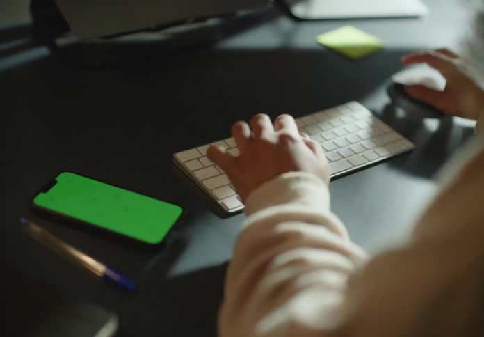 Woman with curly hair and brown sweater types on keyboard and uses smartphone