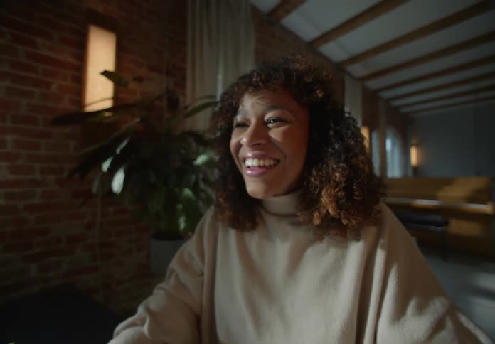 Woman with brown wavy hair beige turtleneck sweater sits at desk looking at laptop and laughing next to houseplant and brick wall in room