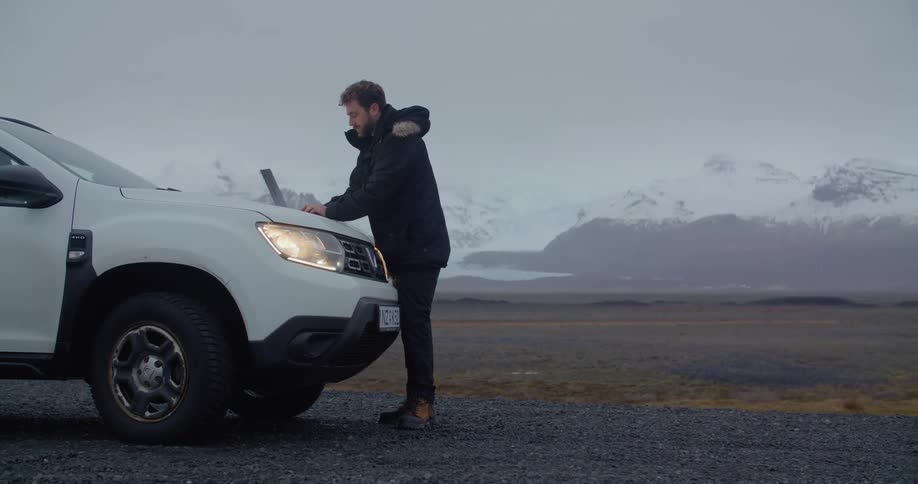 A man wearing a winter coat is typing on a laptop computer sitting on the front of a white automobile and there are snow covered mountains in the distance