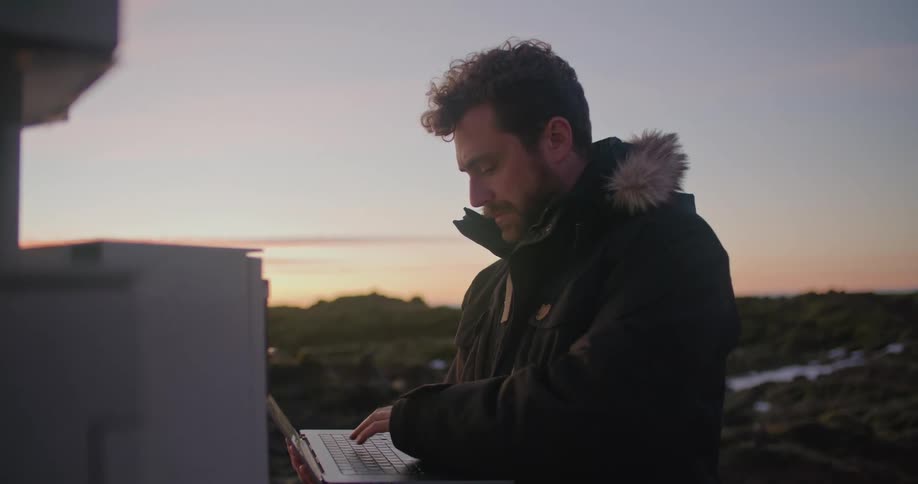A man is standing outside in front of a utility box holding a laptop computer and typing with the other hand