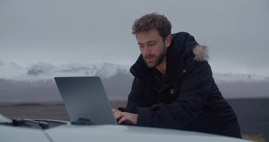 A man wearing a winter coat is typing on a laptop computer sitting on the front of a white automobile and there are snow covered mountains in the distance