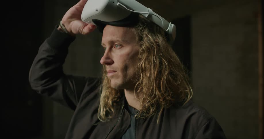 A man with long blond hair wearing a virtual reality headset lifts it off his head