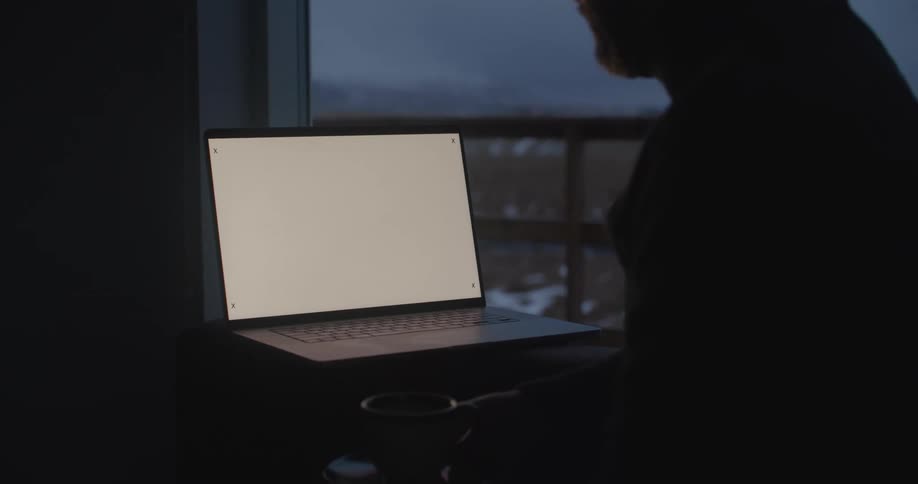 A man is sitting at a table in a dark room looking at a laptop computer near a balcony