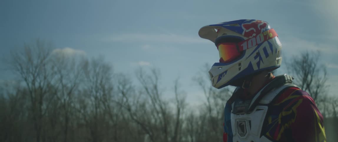 Man wearing colorful motocross protective equipment sitting on a parked motorcycle near a tree line