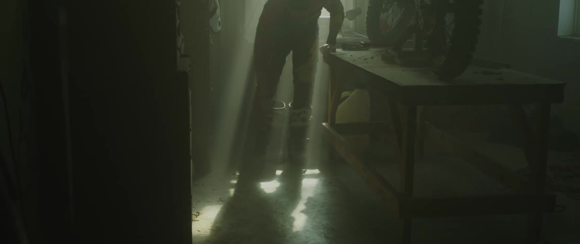 A man stands beside a table with a motorcycle on it and puts boots on