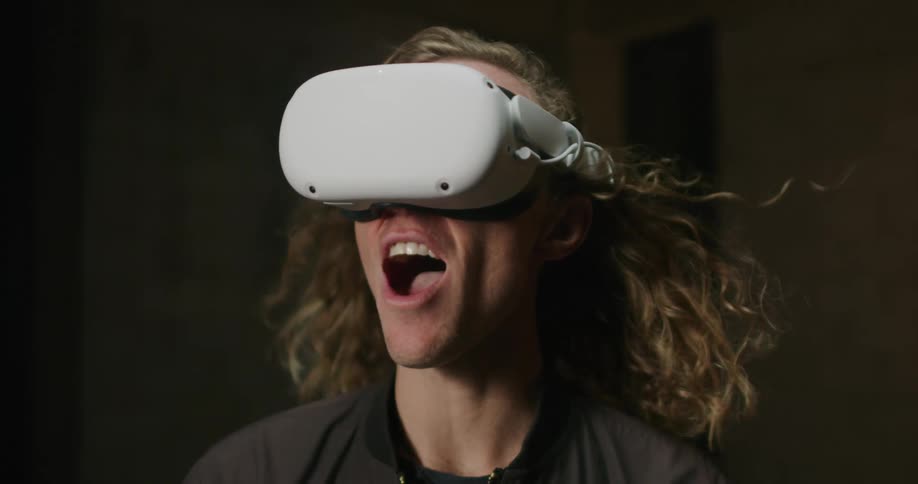 A man with long blond hair wearing a virtual reality headset screams as his hair is blown backwards