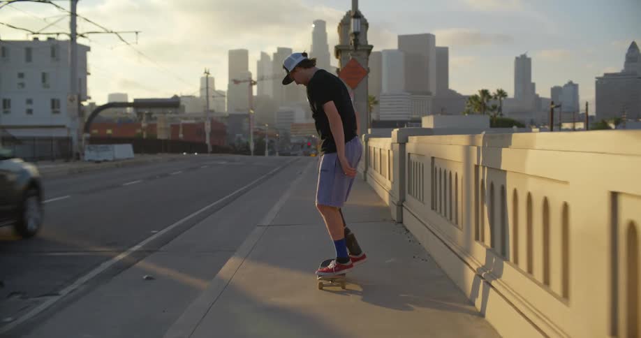 Young man riding on a skateboard over a bridge in front of the city skyline at sunset