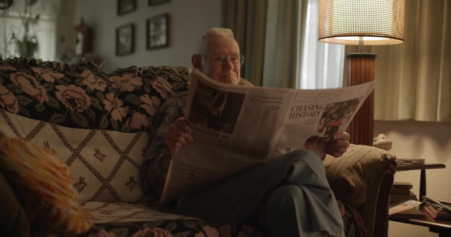 Elderly sad man with glasses shirt and trousers sitting on couch reads newspaper