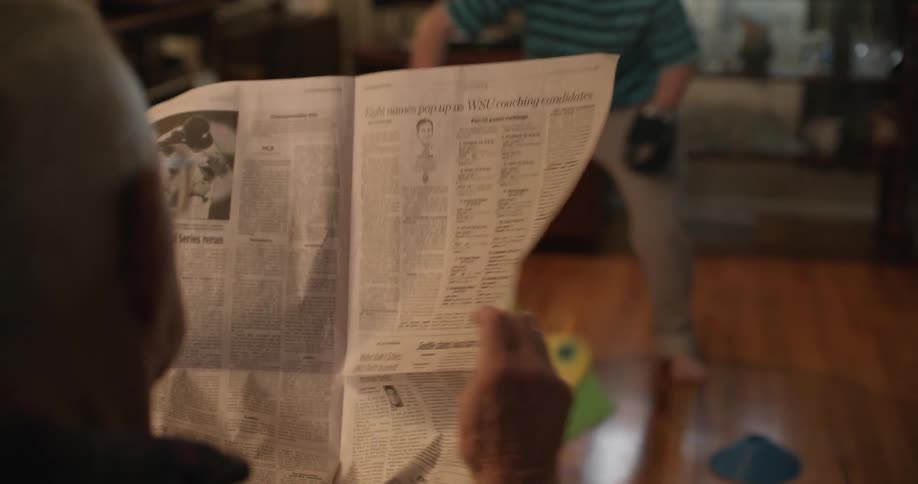 A man reads a newspaper while a girl plays hockey indoors with a broom