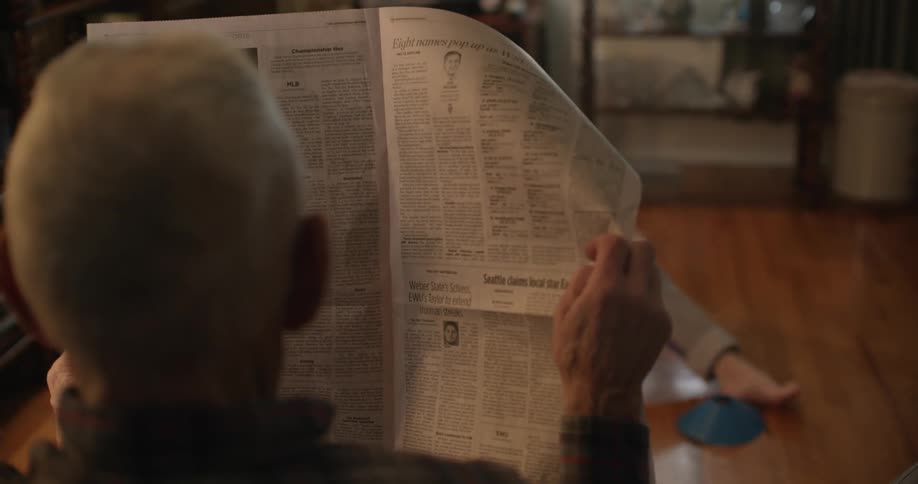 A girl sits on a floor in a home and plays in front of an elderly man who is reading the newspaper