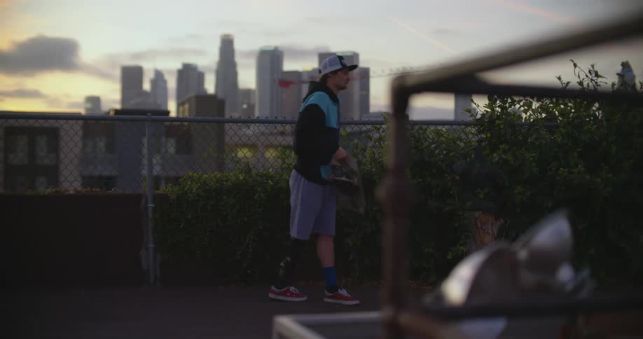A teenage boy is looking over a fence on a rooftop then picks up his skateboard and walks away