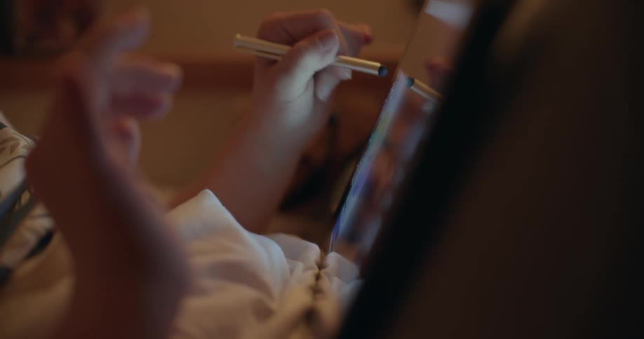 Young girl drawing with a stylus on a tablet screen while resting over the bed at home