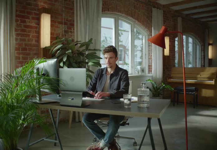 Man with goatee beard dark blue jacket and white shirt and jeans sits at desk typing on dekstop keyboard next to houseplant and brick wall in room