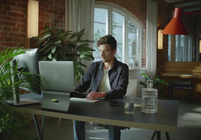 A man is sitting at a table typing on a keyboard and he picks up his cell phone to make a call
