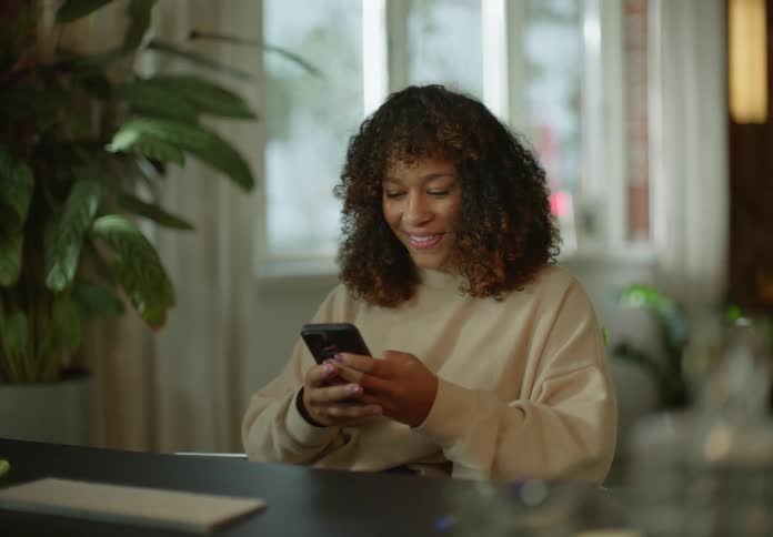 Woman with curly hair and brown sweater sits in armchair and uses smartphone in living room at home