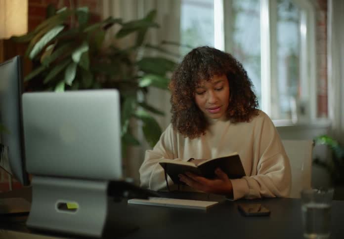 A woman is sitting at a desk looking through a book and talking to someone on the computer