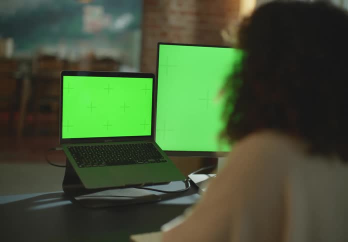 Woman with brown curly hair and beige turtleneck sweater types on computer keyboard looking at laptop and desktop with green screens in room