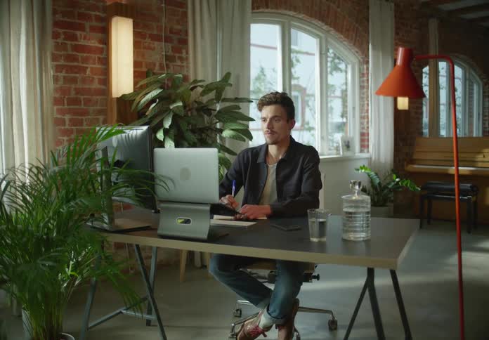 Man with dark blue jacket white shirt and jeans sits at desk uses laptop while writing down in notebook in office