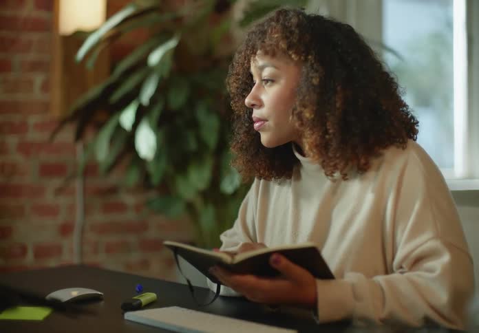 Woman with brown curly hair and beige turtleneck sweater writes down in notebook looking at laptop next to brick wall in room