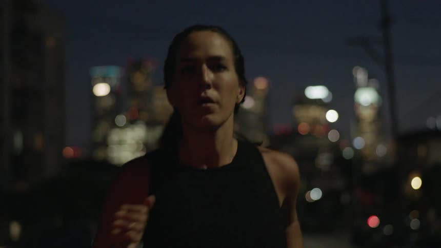 A woman is running in the street at night with a city behind her