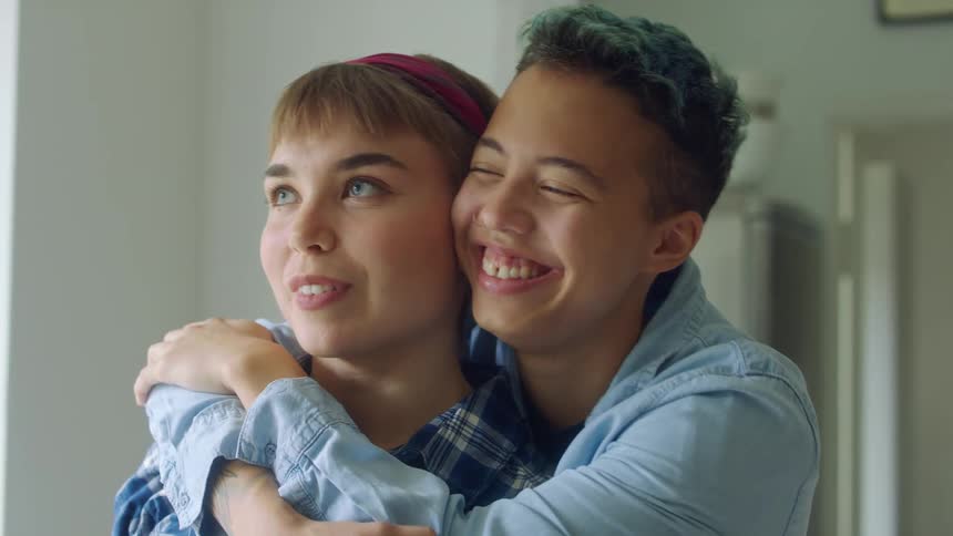 A teenage boy stands behind a girl with his arms hugging her around her neck as they laugh and talk