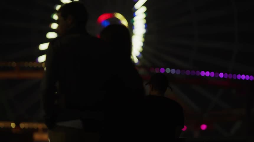 People walk past a flashing ferris wheel at night
