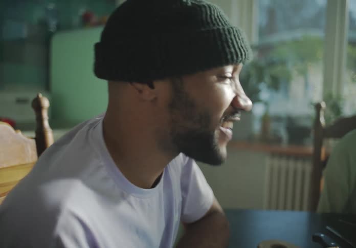 Man with black beanie beard and white t shirt sits at dining table talking to friends in the kitchen