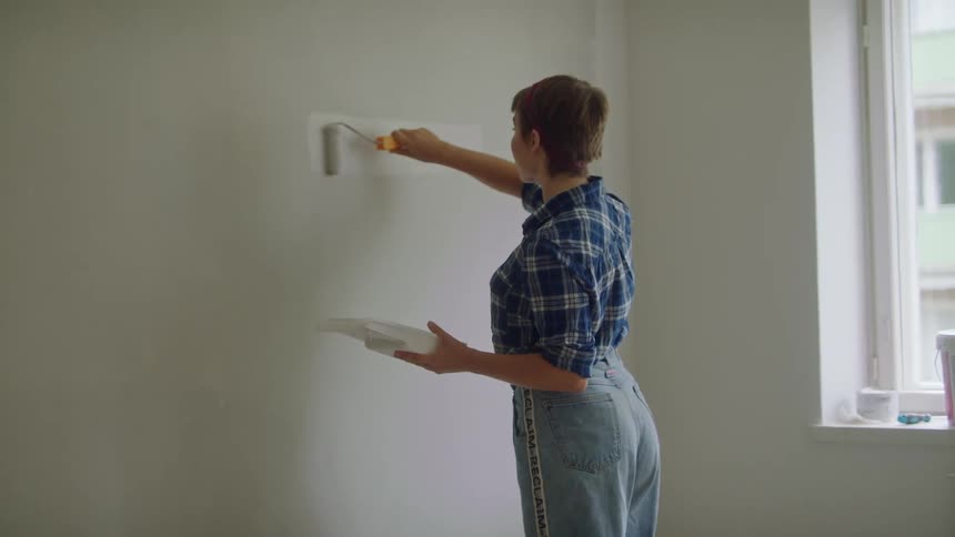Woman with brown short hair blue plaid shirt and jeans paints wall white in room