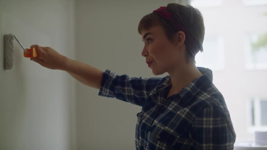 Woman with short hair and blue plaid shirt paints wall white in room