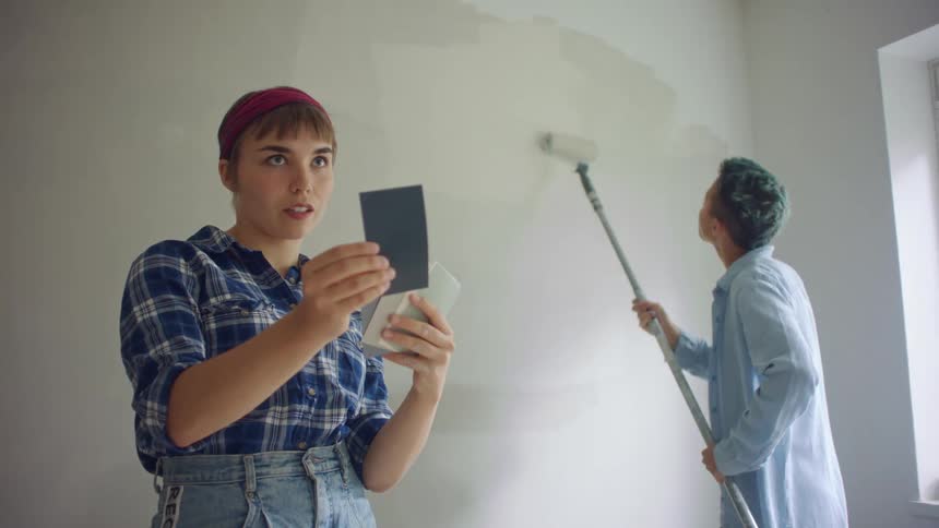 A woman is painting as the other woman looks at paint colors