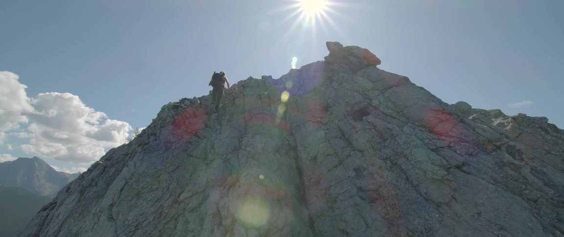 A mountain climber is near the top of a mountain as the sun shines brightly in the sky