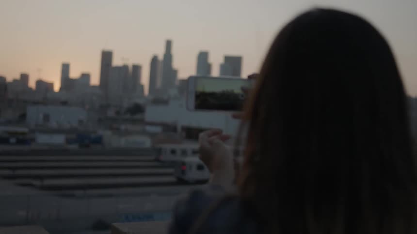 A woman is holding a camera in front of her taking a photograph at sunset