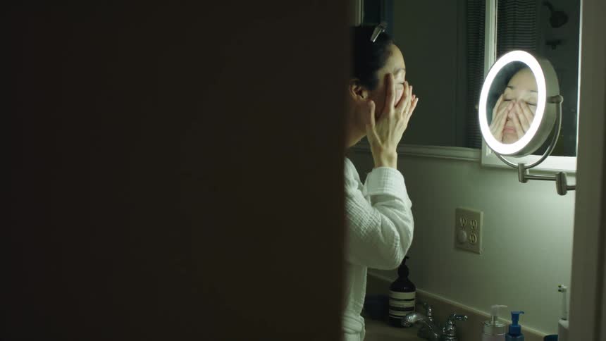 Woman rub her face while standing in bathroom looking into makeup mirror