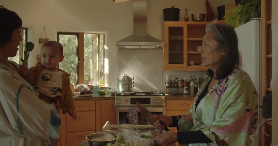 Woman holding a baby on her arms stands in front of her mother cooking in the kitchen at home
